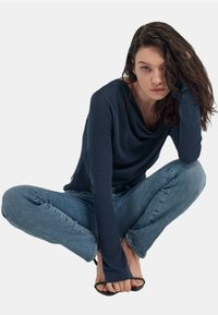 Maglietta a maniche lunghe blu navy con scollo drappeggiato, abbinata a jeans azzurri chiari. Il modello è accovacciato, tenendo un piede. Sandali neri flat visibili.
