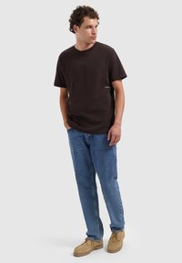 Bruine katoenen T-shirt met korte mouwen, ronde hals en een klein wit logo, gecombineerd met lichtblauwe jeans en beige schoenen.