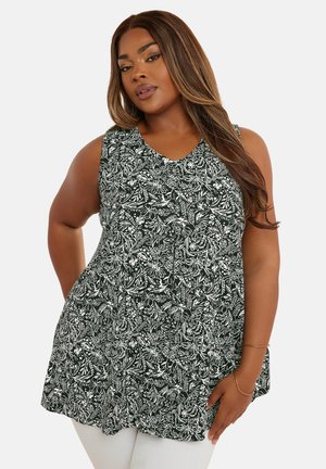 Femme portant un haut tunique sans manches à motif floral noir et blanc avec un pantalon blanc, posant avec une main sur la hanche et l'autre détendue.