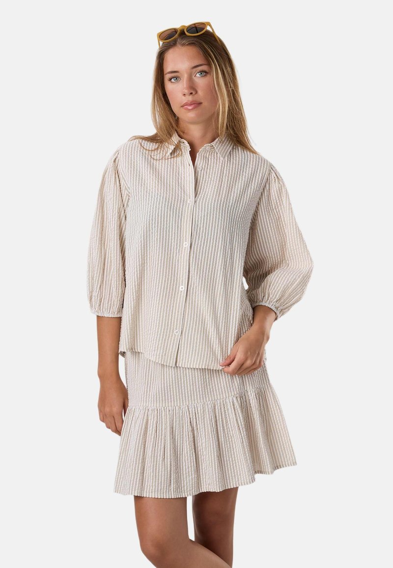Femme portant un chemisier beige à rayures blanches avec manches 3/4 bouffantes et jupe évasée assortie, des lunettes de soleil posées sur la tête.