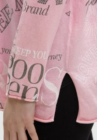 Hand ruht nahe dem Saum eines rosa Langarmshirts mit schwarz-weißem Text, kombiniert mit schwarzen Hosen.