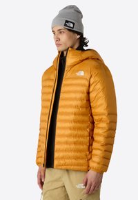 Pufová bunda v jasně oranžové barvě, s prošívanou texturou, kapucí a zipovým zapínáním. Nosená přes černý top s béžovými kalhotami.