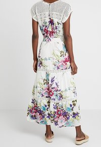 Maxi kjole med hvid blomsterprint, der har lilla og blå roser, hæklede detaljer på livet og lagdelt design. Blødt, flydende stof.