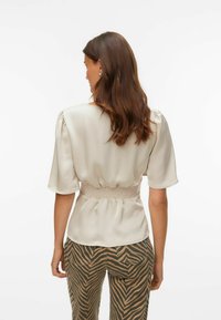 Løs, kremfarget bluse med smockdetalj rundt midjen og korte ermer, kombinert med brune og beige stripete bukser.