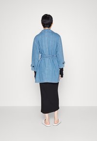 Džínová bunda s páskem v pase, dlouhými rukávy a manžetami na knoflíky, doplněná černými šaty dlouhými ke kotníkům a bílými nazouvacími botami.