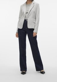 Grijze blazer met een getailleerd ontwerp, gedragen over een lichtgrijs topje, gecombineerd met donkerblauwe broek met wijde pijpen en zwarte puntige platte schoenen.