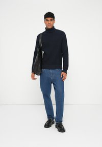 Pull en laine bleu marine à col roulé, jeans bleu décontractés et chaussures noires en cuir à enfiler. Modèle tenant un sac fourre-tout noir, se tenant devant un fond uni.