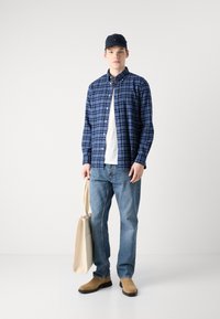 Camisa de cuadros azul sobre una camiseta blanca, jeans de mezclilla azules, zapatos de ante beige y un bolso de tela beige en un entorno minimalista.