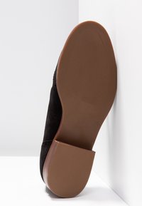Semelle d'une chaussure en daim noir avec une semelle en caoutchouc marron et un talon bloc, montrant la texture antidérapante et le nom de la marque embossé sur la zone du talon.