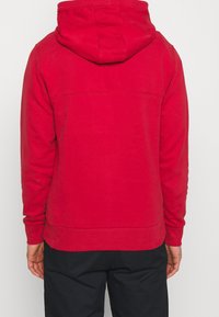 Sudadera roja con capucha, mangas largas, puños y dobladillo de canalé. Textura suave, detalles de costura mínimos en la parte superior de la espalda.