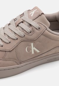 Beige lærersneakers har en tykk såle, teksturert overflate og en subtil CK-logo på siden. Snøringene er lys grå og flate.