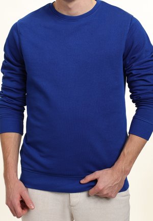 Homme portant un pull à col rond à manches longues bleu uni et un pantalon beige clair, ajustant l'ourlet du pull d'une main.