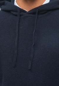 Tamsiai mėlynas purus džemperis su gobtuvu, pasižymintis minkšta tekstūra, turintis virvutes gobtuve ir išryškintus siuvimo detales kaklo bei pečių siūlėse.