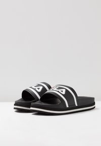Chanclas negras con acentos blancos, que presentan una tira ancha y un logo. Material suave, suela plana con un sutil detalle de raya blanca.