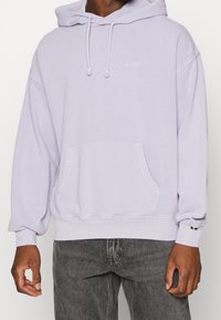 Sweat à capuche lavande avec une poche kangourou, une capuche à cordon de serrage et des poignets côtelés. Présente un logo subtil sur la poitrine et une coupe décontractée.