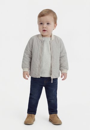 Bambino piccolo in piedi, indossa una giacca a righe beige, una maglia grigio chiaro, jeans blu scuro e stivali in camoscio color cuoio, su sfondo bianco uniforme.