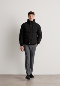 Jachetă puffer neagră cu închidere prin butoane în față și guler înalt, combinată cu pantaloni gri croiți și pantofi negri, stând pe un fundal deschis.