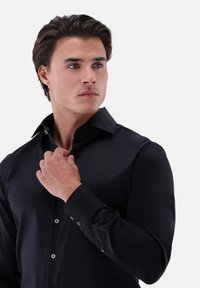 Schwarzes Hemd aus glattem Stoff. Mit spitzem Kragen, Manschetten mit Knöpfen und kleinen Knöpfen an der Vorderseite. Figurbetontes Design.