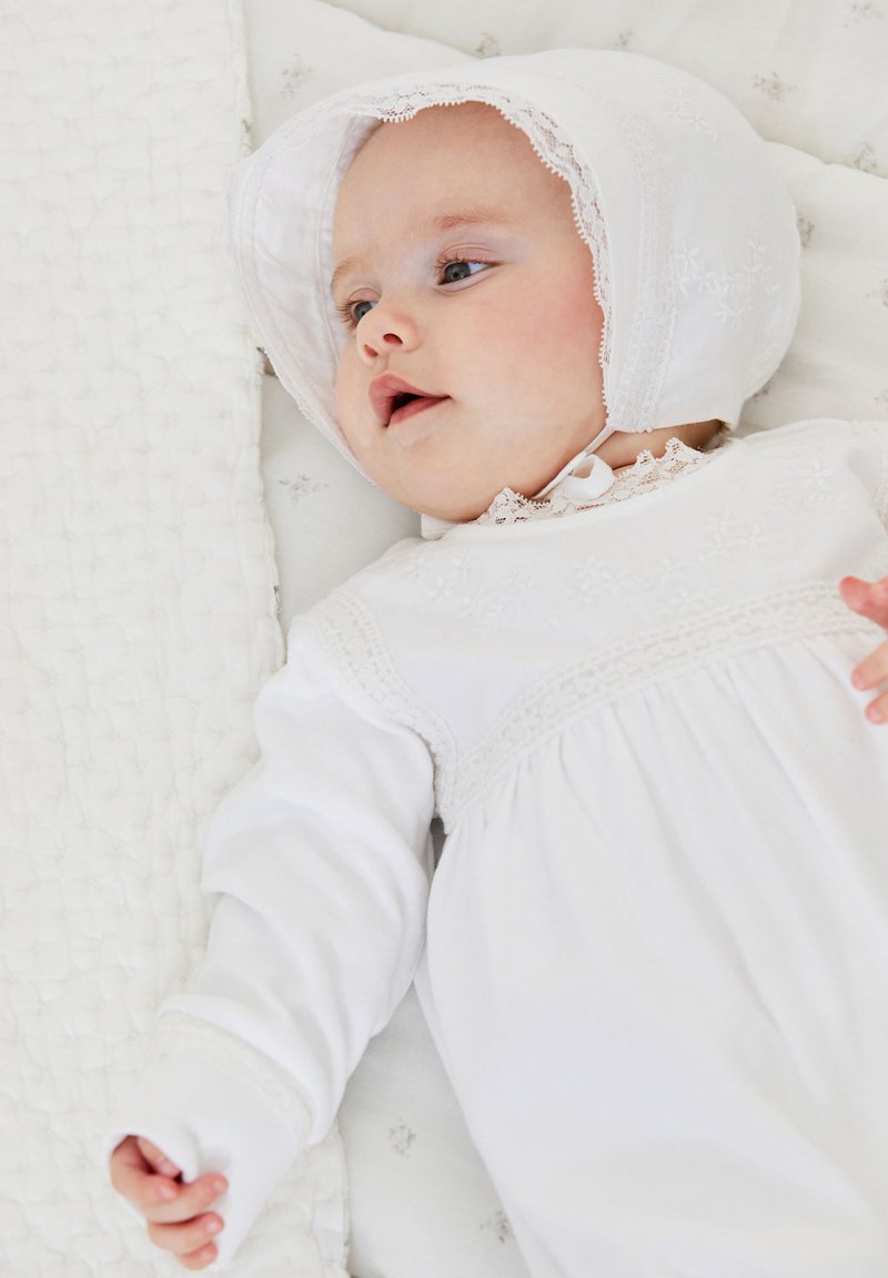 Vit babydress med broderade blommönster, spetskant och en matchande mössa. Bebisen ligger på en strukturerad ljus filt.