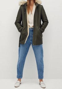 Oliwkowa parka z kapturem z sztucznego futra, zapinana na zamek, z dwiema przednimi kieszeniami, noszona na beżowym swetrze i niebieskich dżinsach z wysokim stanem.