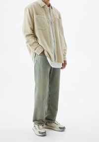 Chemise en velours côtelé beige à boutons avec deux poches poitrine, portée par-dessus un t-shirt blanc. Jean baggy bleu clair et baskets grises, blanches et marine.