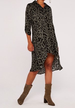 Femme portant une robe imprimé léopard verte et noire avec des manches retroussées et des bottes mi-mollet en daim marron, debout sur un fond uni.