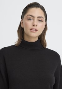 Fiatal nő barna, hátrasimított hajjal, fekete garbót visel, semleges arckifejezéssel egyenesen előre néz.