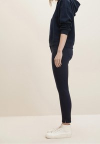 Navy hoodie with a kangaroo pocket, relaxed fit, and ribbed cuffs paired with slim-fit navy pants and white high-top sneakers.