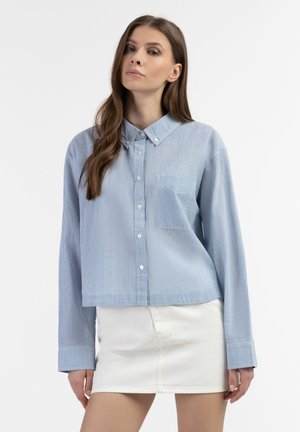 Camicia a maniche lunghe, blu chiaro, con bottoni e taschino, caratterizzata da un sottile motivo a righe e colletto. Abbinata a una gonna bianca.