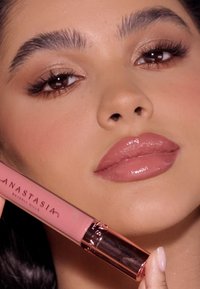 Płynna pomadka w przezroczystej tubie z różowym złotym wieczkiem. Odcień to delikatny róż, z błyszczącym wykończeniem, trzymana blisko gładkich, lekko zabarwionych warg.