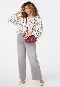 Femme aux cheveux bouclés portant un chemisier blanc à cœurs noirs, un jean large gris, des ballerines beiges, et tenant un sac bandoulière bordeaux.