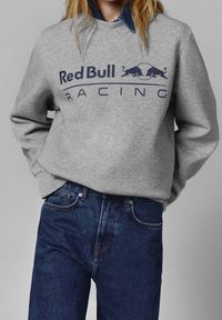 Szara bluza z nadrukiem "Red Bull Racing" w kolorze granatowym, z okrągłym dekoltem i luźnym krojem, zestawiona z ciemnoniebieskimi jeansami.