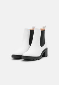 Bottines blanches en cuir avec des panneaux élastiques noirs, une semelle en caoutchouc et un talon block. Présente une languette à l'arrière.