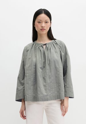 Junge Frau mit glattem dunklem Haar, die eine lockere, hellgraue Bluse mit langen Ärmeln und gebundenem Ausschnitt sowie offwhite Hose trägt.