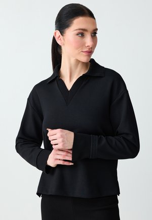 Kobieta o ciemnych włosach związanych w kucyk, ubrana w czarny sweter z długim rękawem i dekoltem w serek z kołnierzykiem, patrząca w prawo z delikatnie splecionymi dłońmi.