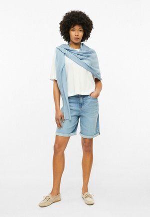 Femme debout face à l’avant, portant une chemise blanche, un short en jean, un pull bleu clair posé sur les épaules et des chaussures bateau beiges, la main dans la poche.