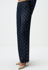 Marineblauwe broeken met een gladde textuur, voorzien van een geometrisch patroon in lichte kleuren. Gekleed met nude high heels.