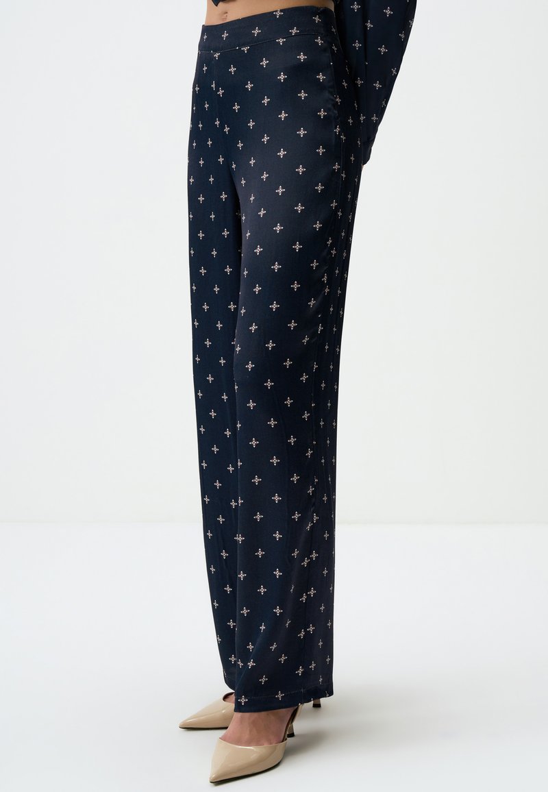 Marineblauwe broeken met een gladde textuur, voorzien van een geometrisch patroon in lichte kleuren. Gekleed met nude high heels.