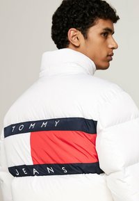 Giacca a vento bianca con colletto alto; presenta una striscia orizzontale blu marino e rossa con la scritta "TOMMY JEANS" sul retro. Texture liscia.