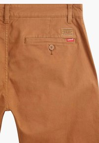 Bruna bomullschinos med en knäppt bakficka, ett vävt etikettmärke och en röd varumärkesetikett, med en slät och hållbar tygtextur.