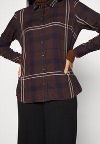 Femme portant une chemise à carreaux marron à boutons sur un col roulé marron, les mains sur les hanches, assortie à un pantalon noir.