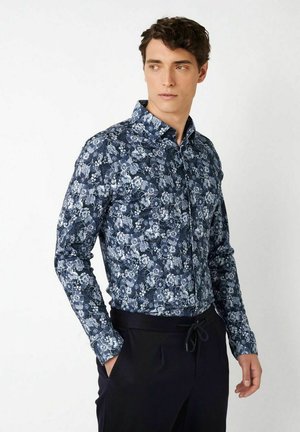 Junger Mann mit einem langärmligen, dunkelblauen Hemd mit Blumenmuster und schwarzer Hose, die Hände in den Taschen, schaut zur Seite.