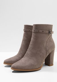 Šedé kotníkové boty z broušené kůže s vysokým blokovým podpatkem, špičatou špičkou a páskem kolem kotníku zdobeným dekorativními nýty a kovovou sponou.