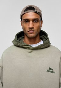 Beige oversized Kapuzenpullover mit einem olivgrünen Innenfutter. Hat "Stay Moment" in Grün gestickt. Das Model trägt eine hellbraune Mütze.