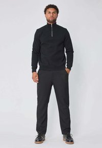 Sweater noir à col zippé avec des manches longues, associé à un pantalon noir. La tenue comprend des chaussures noires avec une semelle texturée.