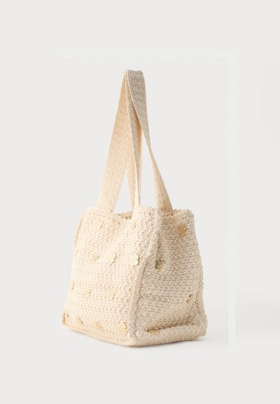 Τσάντα ώμου κρεμ χρώματος, πλεκτή με βελονάκι, με φαρδιές λαβές και μικρές χρυσές διακοσμητικές λουλούδια σε μονόχρωμο φόντο.