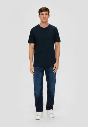 Navy blue cotton t-shirt with a round neckline, paired with dark denim jeans. Model wears white sneakers, standing against a plain background.