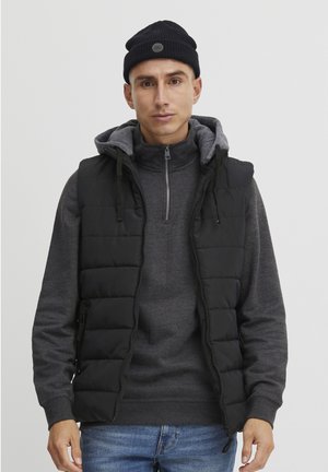 Black puffy vest over a gray hoodie with a zippered collar, paired with blue jeans. The model wears a black ribbed beanie.