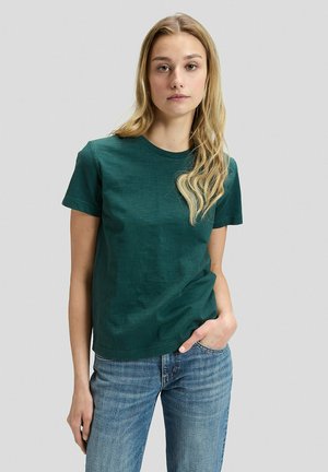 Młoda kobieta z długimi blond włosami ma na sobie ciemnozielony t-shirt i niebieskie dżinsy, stoi z jedną ręką w kieszeni na tle jednolitego tła.