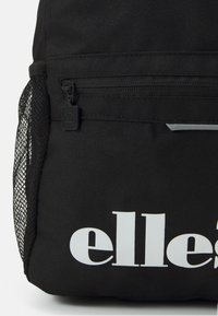Fekete szövet hátizsák textúrált felülettel, hálós oldalsó zsebével és fehér logójával az elülső zseben. Cipzáras rekesz található benne.